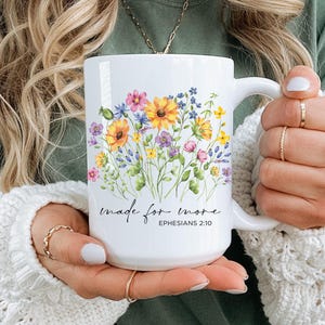 May include: White ceramic mug featuring a floral design with yellow, purple, and pink flowers. The mug has the text "made for more" and "Ephesians 2:10" printed on it. The mug has a large handle.