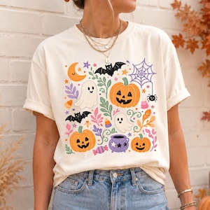 Może przedstawiać: Kremowy t-shirt z kr&oacute;tkim rękawem z kolorowym nadrukiem graficznym o tematyce Halloween, przedstawiającym pomarańczowe dynie, białe duszki, czarne nietoperze, fioletowy kociołek, pajęczynę, p&oacute;łksiężyc oraz r&oacute;żnego rodzaju cukierki.