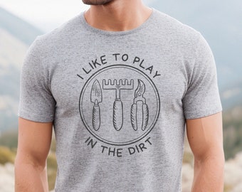 Puede incluir: Una camiseta gris con el texto "Me gusta jugar en la tierra" y un gráfico de herramientas de jardinería.