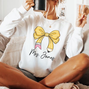 May include: White sweatshirt with a yellow bow and a pencil design. The text "Mrs. Jones" is printed below the bow.