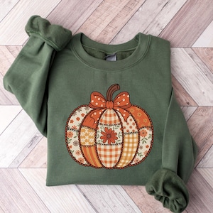 Peut inclure: Sweat-shirt vert avec un motif de citrouille en patchwork. La citrouille a une tige marron et un grand nœud orange à pois blancs. La citrouille est faite de divers motifs floraux et à carreaux orange et jaune.