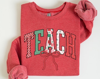 May include: Red sweatshirt with the word "TEACH" in a collegiate style. The letters are decorated with Christmas-themed patterns, including stripes, trees, and snowflakes. A red bow is below the word.