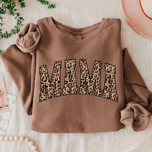 May include: A brown sweatshirt with the word "MAMA" in a leopard print design across the front. The sweatshirt is folded, with the sleeves draped to the side. A hat and greenery are in the background.