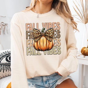 May include: Cream-coloured long-sleeve shirt with a pumpkin graphic and the text "FALL VIBES". The pumpkin has a leopard print bow. The shirt is a fall-themed apparel item.