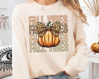 May include: Cream-colored long-sleeve shirt with a pumpkin graphic and the text "FALL VIBES". The pumpkin has a leopard print bow. The shirt is a fall-themed apparel item.