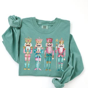 May include: A teal long-sleeve shirt featuring a watercolor illustration of four nutcracker figures. The nutcrackers are in various colors, including pink, teal, and gold, with crowns and ornate details. The shirt has a crew neck and long sleeves.
