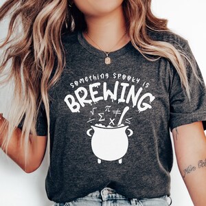 May include: A dark grey t-shirt with a white graphic that says "Something spooky is brewing" with a cauldron and mathematical symbols. The shirt is being worn by a person with long blonde hair.