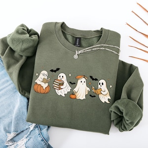 May include: An olive green sweatshirt featuring a graphic of four cartoon ghosts reading books, with pumpkins and bats. A silver necklace is draped on the sweatshirt. The sweatshirt is paired with blue jeans.
