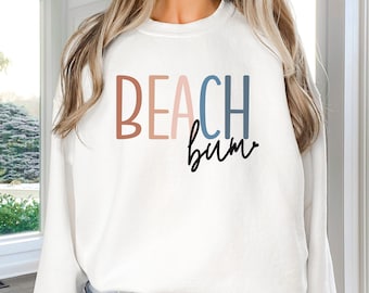 May include: White sweatshirt with the text "BEACH bum" in colorful letters. The letters are in shades of brown, pink, blue, and light blue.