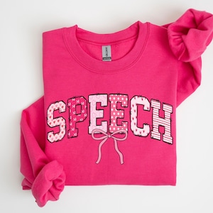 Peut inclure: Un sweat-shirt rose vif avec le mot "SPEECH" en lettres décoratives sur le devant. Les lettres sont dans des tons roses avec des motifs de cœurs, de pois et de rayures. Un nœud rose est centré sous le mot.