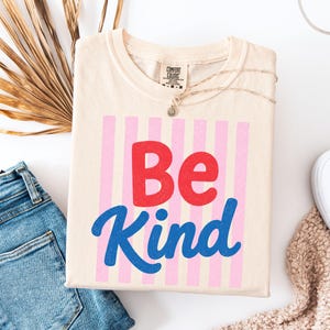 May include: A cream-colored t-shirt featuring a graphic with vertical light pink stripes. The text "Be" is printed in bold red letters, and the word "Kind" is printed below in a blue script font. The shirt has a Comfort Colors brand label.