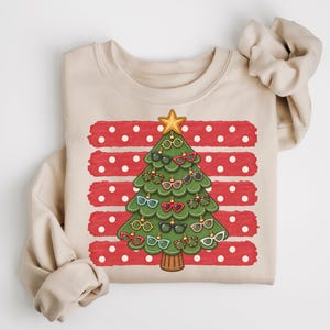 May include: A beige crewneck sweatshirt featuring a graphic of a green Christmas tree decorated with various colourful eyeglasses as ornaments. The tree is set against a background of horizontal red brushstroke stripes with white polka dots.