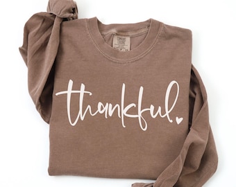 Puede incluir: Una camiseta marrón de manga larga con la palabra "thankful" en escritura blanca. La camiseta tiene cuello redondo y un pequeño corazón al final de la palabra. La camiseta está hecha de un material suave.