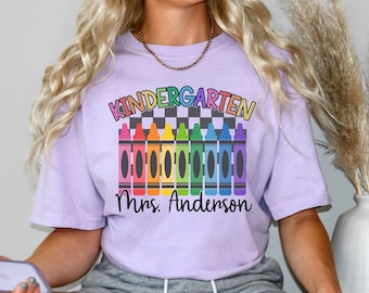 Może przedstawiać: Fioletowy t-shirt z napisem "Kindergarten" w kolorach tęczy i rzędem tęczowych kredek. Pod kredkami znajduje się napis "Mrs. Anderson".