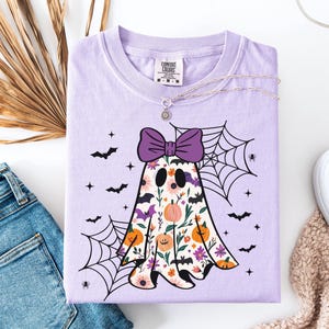May include: A lilac Comfort Colors t-shirt with a floral ghost design, a purple bow, bats, and spiderwebs. The ghost has a floral pattern with pumpkins and flowers. A silver necklace is on the shirt.