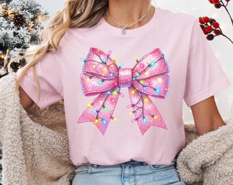 May include: A pink t-shirt with a large pink bow decorated with colorful Christmas lights. The bow is centered on the shirt and is the main focus of the design.