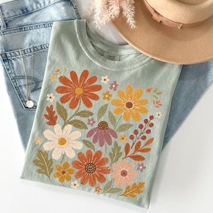 May include: A sage green t-shirt featuring a colorful graphic print of various flowers including orange, yellow, white, and purple daisies, along with green leaves and small red berries. The shirt is layered with light blue denim jeans and a tan hat.