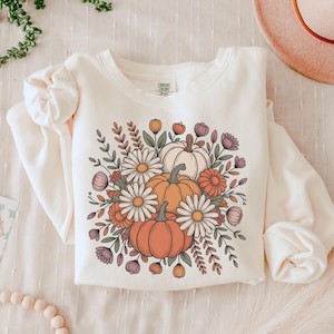 May include: Cream-colored sweatshirt featuring a floral design with pumpkins, daisies, and fall foliage in orange, white, and purple. The long-sleeved top has a crew neck and a relaxed fit, perfect for autumn.