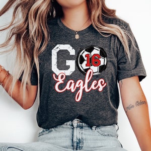 May include: A dark grey t-shirt with a white distressed "GO" and "Eagles" in red script. A black and white football with the number "16" in red is centred between the words.