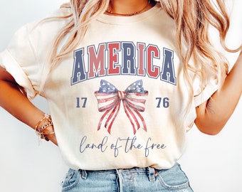 May include: A cream-colored t-shirt with the text "AMERICA" in blue and red letters. The shirt also features a red, white, and blue bow with the numbers "17" and "76" on either side. The text "land of the free" is printed below the bow.