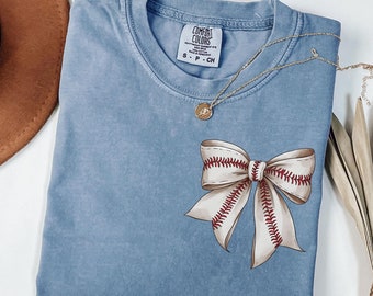 May include: A light blue t-shirt with a white bow made of baseball stitching. The bow has red stitching and is centered on the shirt. The shirt has a tag that says "Comfort Colors" and "S - P - CH".