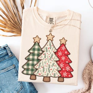May include: A cream-colored t-shirt featuring a graphic of three stylized Christmas trees. The left tree has a green and white buffalo check pattern, the center tree has a botanical print, and the right tree is red with white snowflakes.