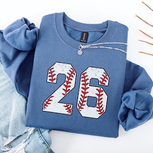 May include: A blue sweatshirt with a white baseball number 26 on the front. The number is outlined in red and has a distressed look. The sweatshirt is laid flat on a white surface.