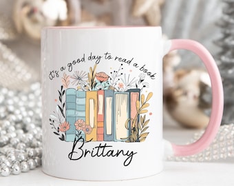 May include: White ceramic mug with a pink rim and handle. It features a colourful illustration of stacked books with flowers and leaves. The text "It's a good day to read a book" is printed on the mug, along with the name "Brittany".