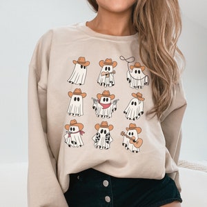 May include: A beige sweatshirt with a graphic print of nine cartoon ghosts wearing cowboy hats. The ghosts are in various poses, including playing a guitar, holding a lasso, and pointing a gun.