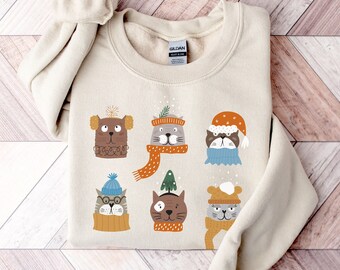 Peut inclure: Un sweat-shirt crème avec un motif fantaisiste de chats de dessins animés portant des bonnets et des écharpes d'hiver. Les chats sont représentés dans différents coloris et styles, avec des flocons de neige. Le sweat-shirt est plié sur une surface en bois.