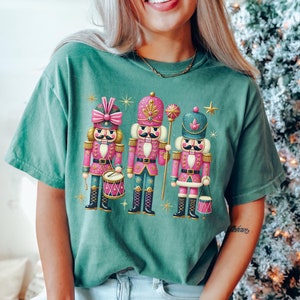 May include: A green t-shirt featuring a festive design with three pink and gold nutcracker figurines. The nutcrackers are holding musical instruments and are surrounded by gold stars.