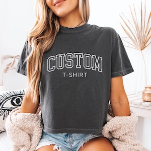 May include: A woman wearing a dark gray cropped t-shirt with the text "CUSTOM T-SHIRT" in white. She is wearing denim shorts and a white fuzzy cardigan.