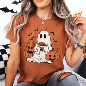 May include: Rust-colored t-shirt featuring a Halloween-themed graphic. The design includes a cartoon ghost holding a coffee cup, flanked by two jack-o'-lanterns and bats. The shirt has short sleeves and a relaxed fit, perfect for the fall season.