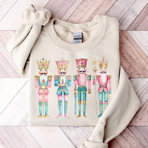 May include: A beige sweatshirt with a watercolor illustration of four nutcracker soldiers. The nutcrackers are wearing gold crowns, pink and blue uniforms, and holding swords and a star.
