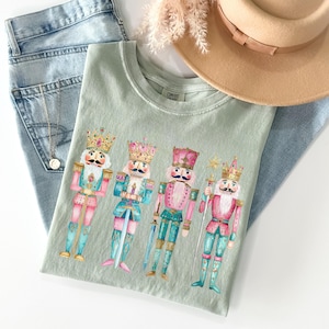 May include: A light green t-shirt with a watercolor design of four nutcracker soldiers. The nutcrackers are wearing colorful costumes and holding swords and other festive accessories.