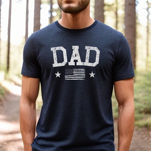 May include: A navy blue t-shirt with a white distressed graphic of the word "DAD" with a small American flag and two stars.