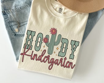 May include: A beige t-shirt with a cactus graphic and the text "Howdy Kindergarten" in a western-style font.