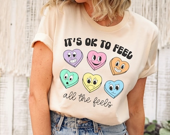 May include: A beige t-shirt with the text "It's ok to feel all the feels" and a graphic of colorful candy hearts with different facial expressions.