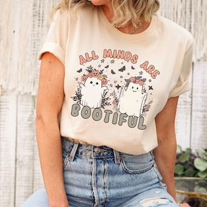 May include: A cream coloured t-shirt with a graphic of two ghosts wearing flower crowns and the text "All Minds Are Bootiful".