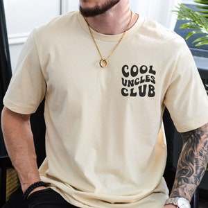 May include: A beige t-shirt with a black graphic that says "Cool Uncles Club" in a retro font. The shirt is being worn by a person with a gold chain necklace.