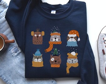 Peut inclure: Un sweat-shirt bleu marine à col rond orné d'un imprimé graphique représentant six visages de chats et de chiens stylisés portant des accessoires d'hiver, notamment des bonnets, des écharpes et des pulls, avec des flocons et un sapin.