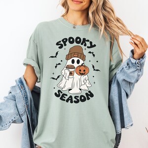 Op de afbeelding: Een lichtgroen T-shirt met een afbeelding van een geest die een beanie draagt en een pompoen en een kopje koffie vasthoudt. De tekst "Spooky Season" is onder de geest gedrukt.