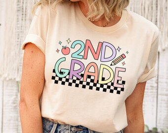 Puede incluir: Una camiseta beige claro con un gráfico colorido que dice "2ND GRADE" con una manzana roja, un lápiz y un patrón de cuadros.