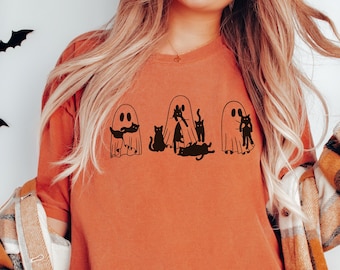 Puede incluir: Una camiseta naranja quemada con un gráfico de tres fantasmas con gatos negros dentro. Los fantasmas están delineados en negro y los gatos son negros.