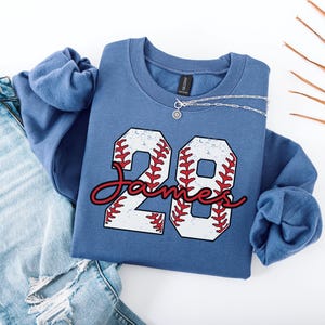 May include: A blue sweatshirt with a white baseball number 28 with red stitching. The name "James" is written in red cursive below the number.