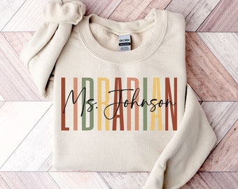 May include: A beige sweatshirt with the text "Librarian" in colorful vertical stripes and the text "Ms. Johnson" in black cursive writing.