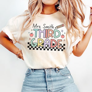 May include: A white t-shirt with the text "Mrs. Smith THIRD GRADE" in colourful letters with a black and white chequered pattern underneath. There are small red apples and stars on either side of the text.