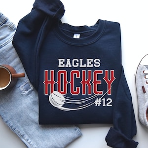 K&ouml;nnte beinhalten: Ein marineblauer Sweatshirt mit dem Text "Eagles Hockey #12" in Rot und Wei&szlig;. Ein Hockeypuck ist in Wei&szlig; mit einem wei&szlig;en Strich dahinter dargestellt.