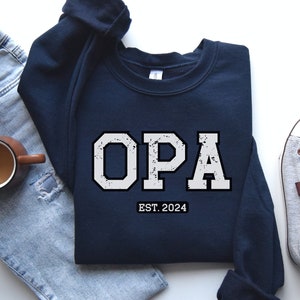 Peut inclure: Sweat-shirt bleu marine avec le mot "OPA" en blanc vieilli de style universitaire. Le texte "EST. 2024" est en dessous. Le sweat-shirt est posé sur une surface blanche.