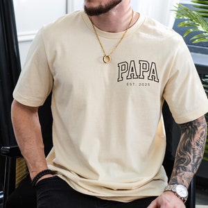 Puede incluir: Una camiseta beige con un gráfico negro que dice "PAPA EST. 2025".